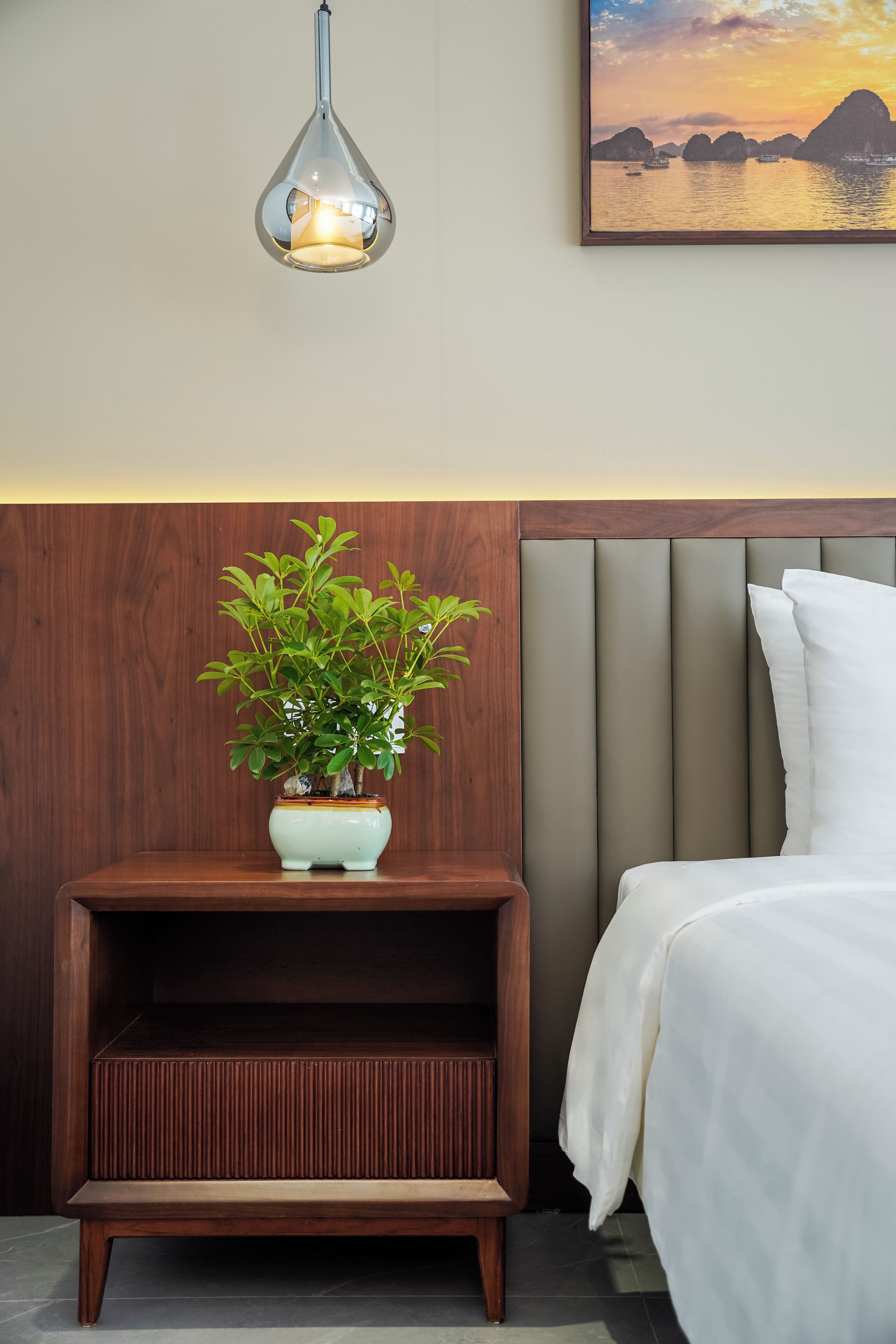 A neatly arranged hotel room with a bed, a bedside table with a potted plant, a wall-mounted light fixture, and a framed picture above the bed.