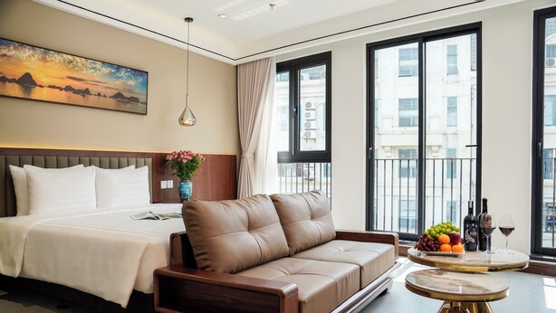 A modern hotel room with a large bed, a leather sofa, a round table with fruit and drinks, and a city view through large windows. - Blubiz Hotel 6 (Hanoi)