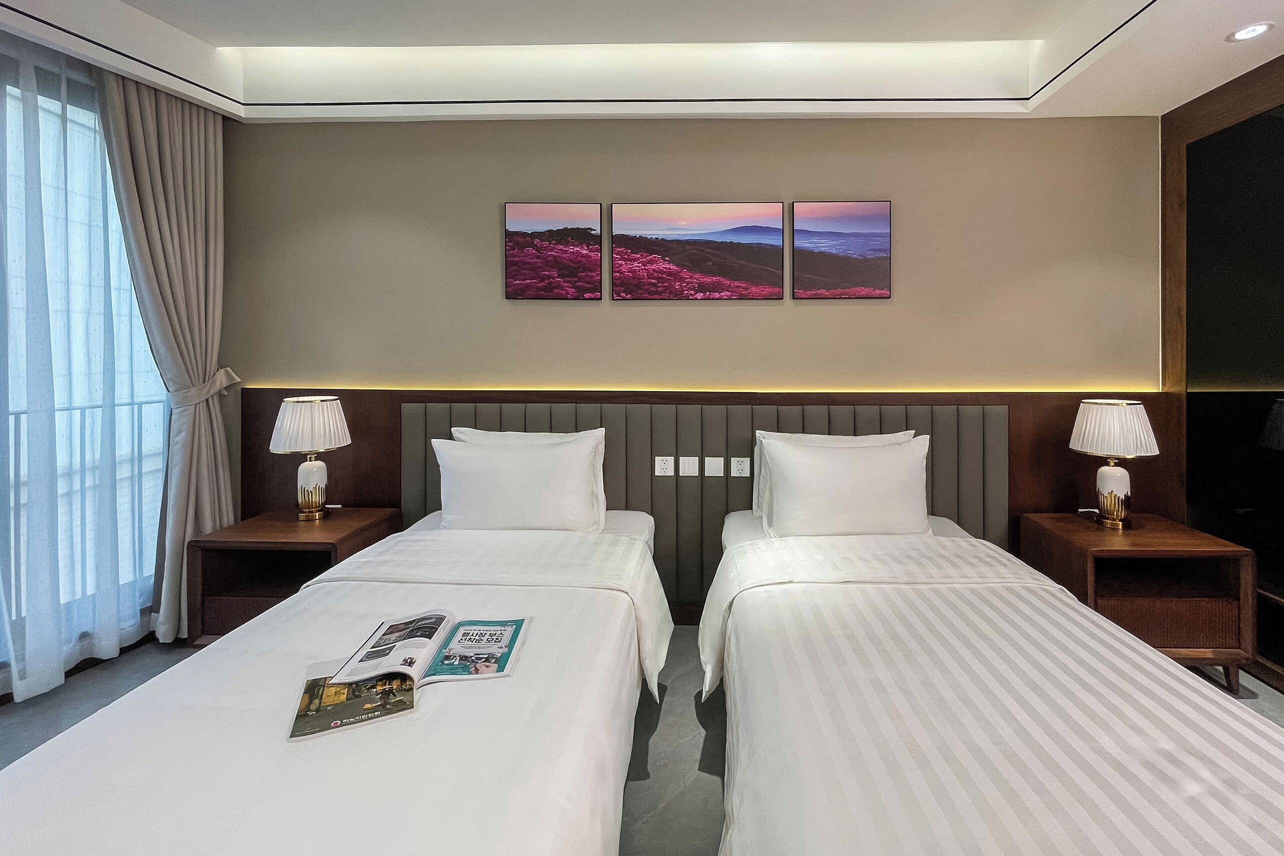 Une chambre d’hôtel avec deux lits, une grande fenêtre avec des rideaux, un tableau accroché au mur et des lampes de chevet.