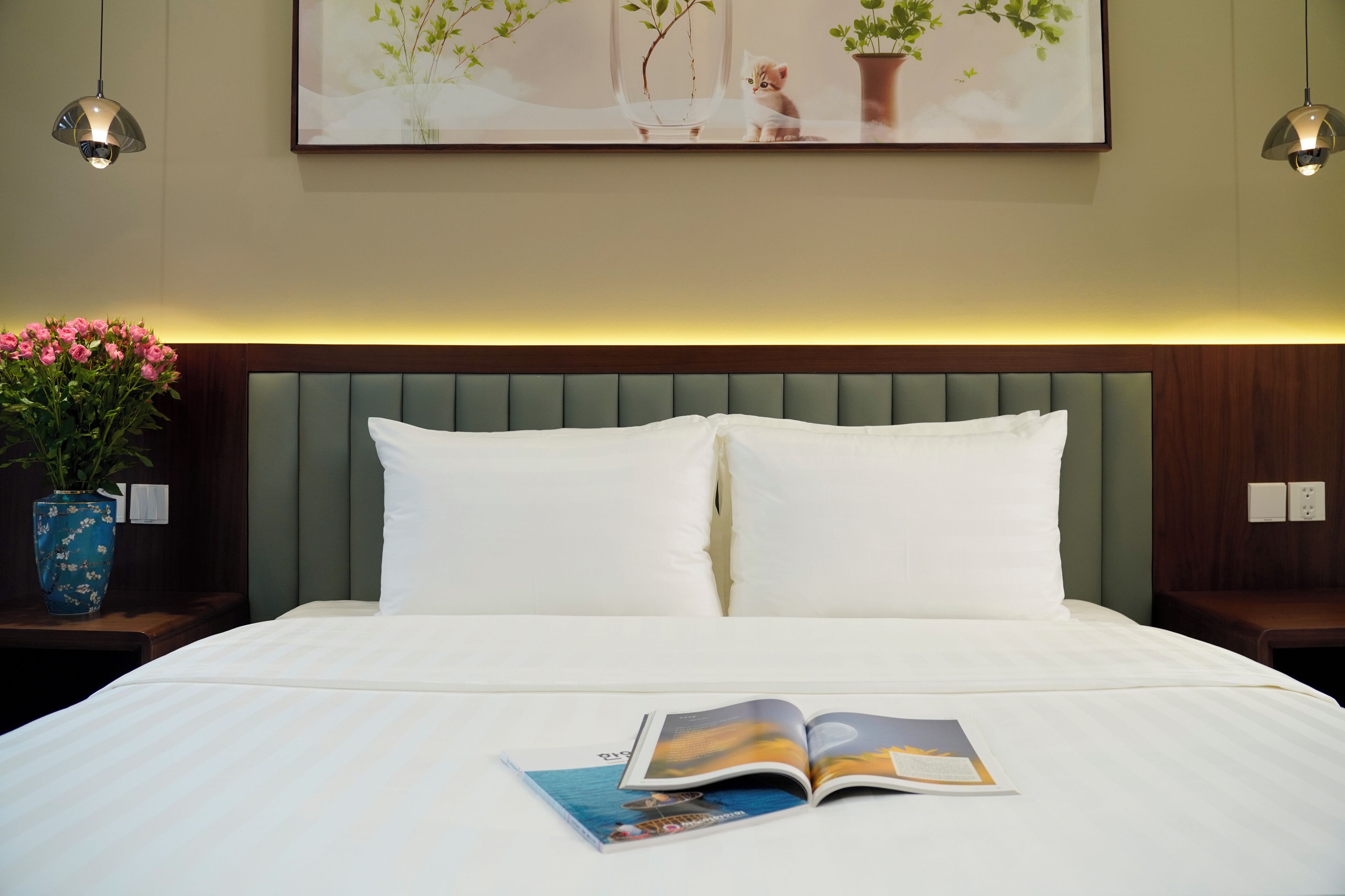 A neatly made bed with a book on it, a vase of flowers, and a painting on the wall.
