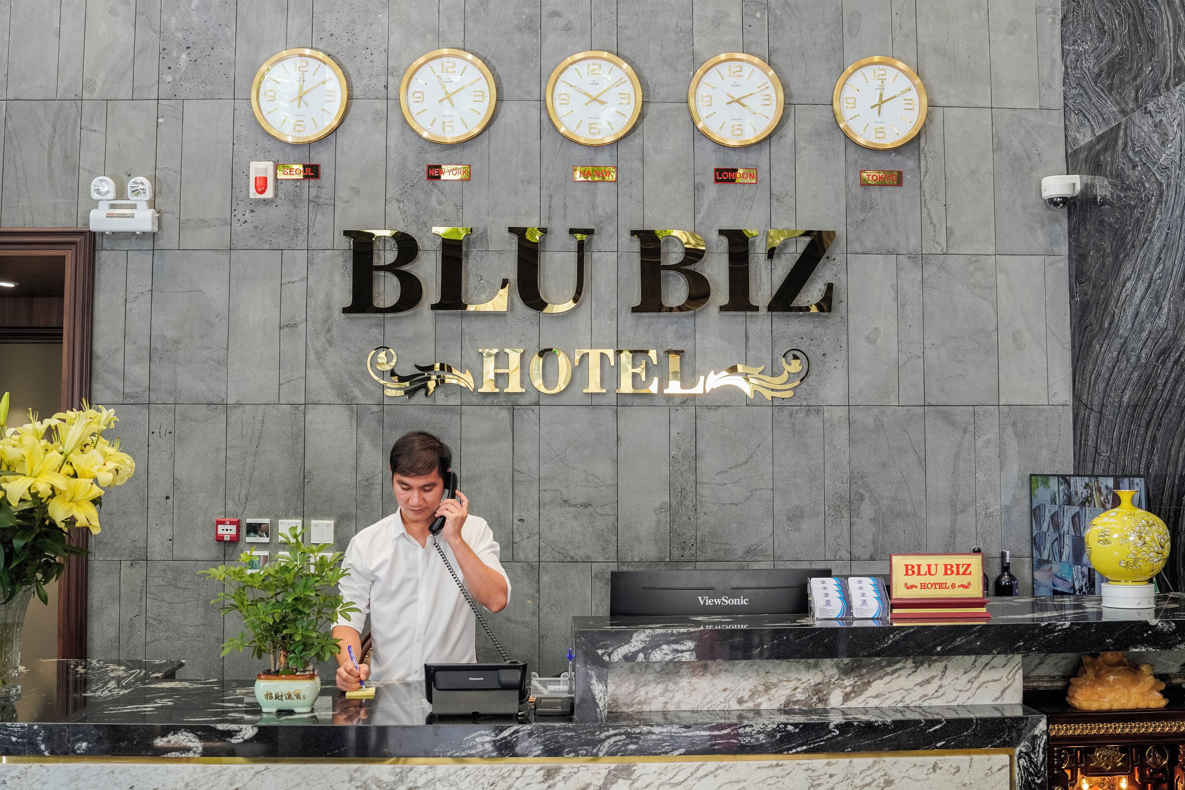 A hotel reception desk with clocks, a BLU BIZ HOTEL sign, a receptionist, and a ViewSonic monitor.