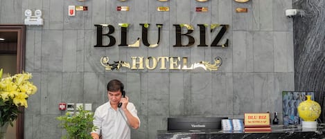 A hotel reception desk with clocks, a BLU BIZ HOTEL sign, a receptionist, and a ViewSonic monitor.