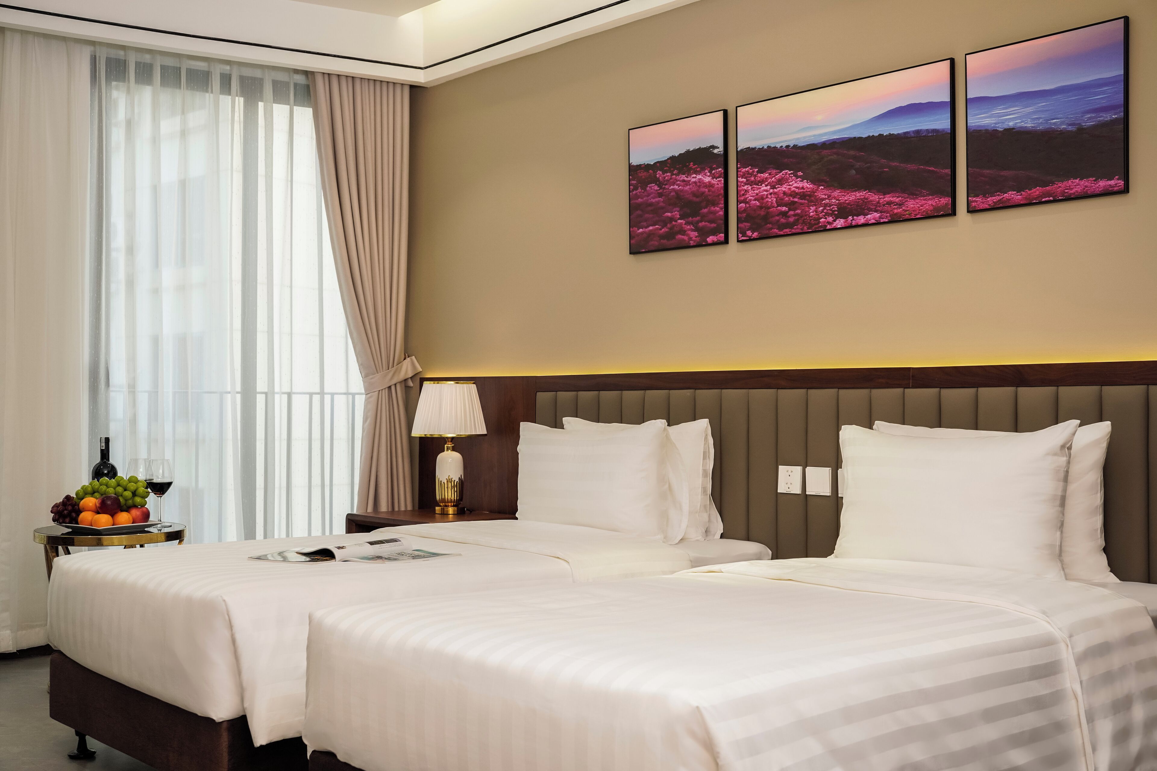 Habitación de hotel con dos camas, una mesita de noche con lámpara, un frutero y una ventana con cortinas.