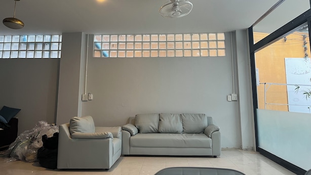 A modern living room with a grey sofa, a small armchair, and a ceiling fan. There is a large window with blinds and a view of the outside. - YP Hostel (Koh Samui)