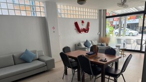 A modern interior with a dining area, sofas, and a large window offering a view of the street. - YP Hostel (Koh Samui)