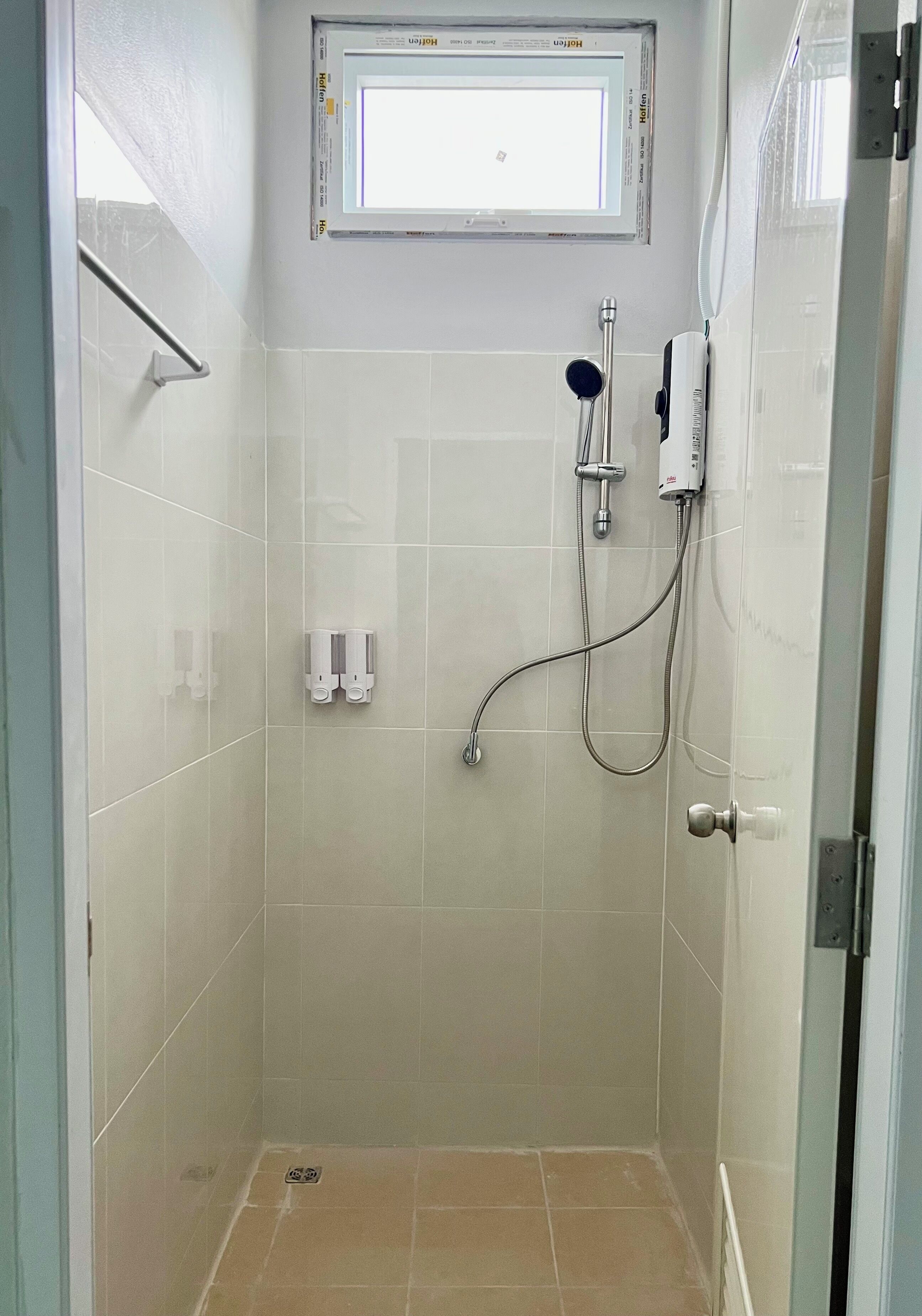 A small, tiled shower room with a handheld showerhead, a wall-mounted faucet, and a window above.