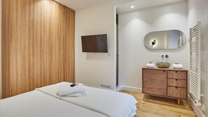 A modern bedroom with a wooden headboard, a flat-screen TV mounted on the wall, a wooden vanity with a mirror, and a white bed with neatly folded towels. - Sumptuous apartment Louvre/Palais Royal (Paris)