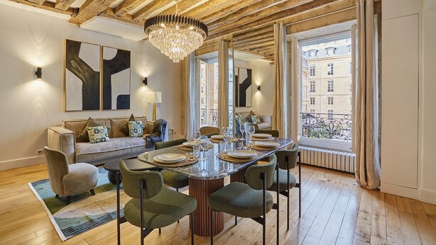 A dining area with a wooden ceiling, a chandelier, a large window with curtains, and a sofa with cushions. - Sumptuous apartment Louvre/Palais Royal (Paris)