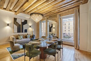 A dining area with a wooden ceiling, a chandelier, a large window with curtains, and a sofa with cushions. - Sumptuous apartment Louvre/Palais Royal (Paris)