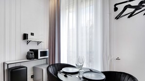 A modern hotel room with a dining area featuring a black table set for two, a small kitchenette, and a large window with sheer curtains. - Superb Suite with AC Beaubourg Le Marais (Paris)