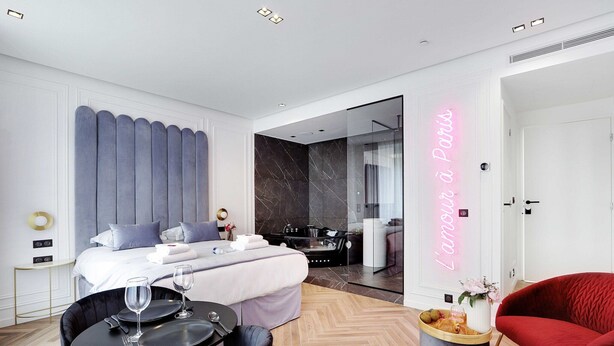 A modern hotel room with a bed, a sitting area with a table and chairs, a glass-enclosed bathroom, and a neon sign on the wall. - Superb Suite with AC Beaubourg Le Marais (Paris)