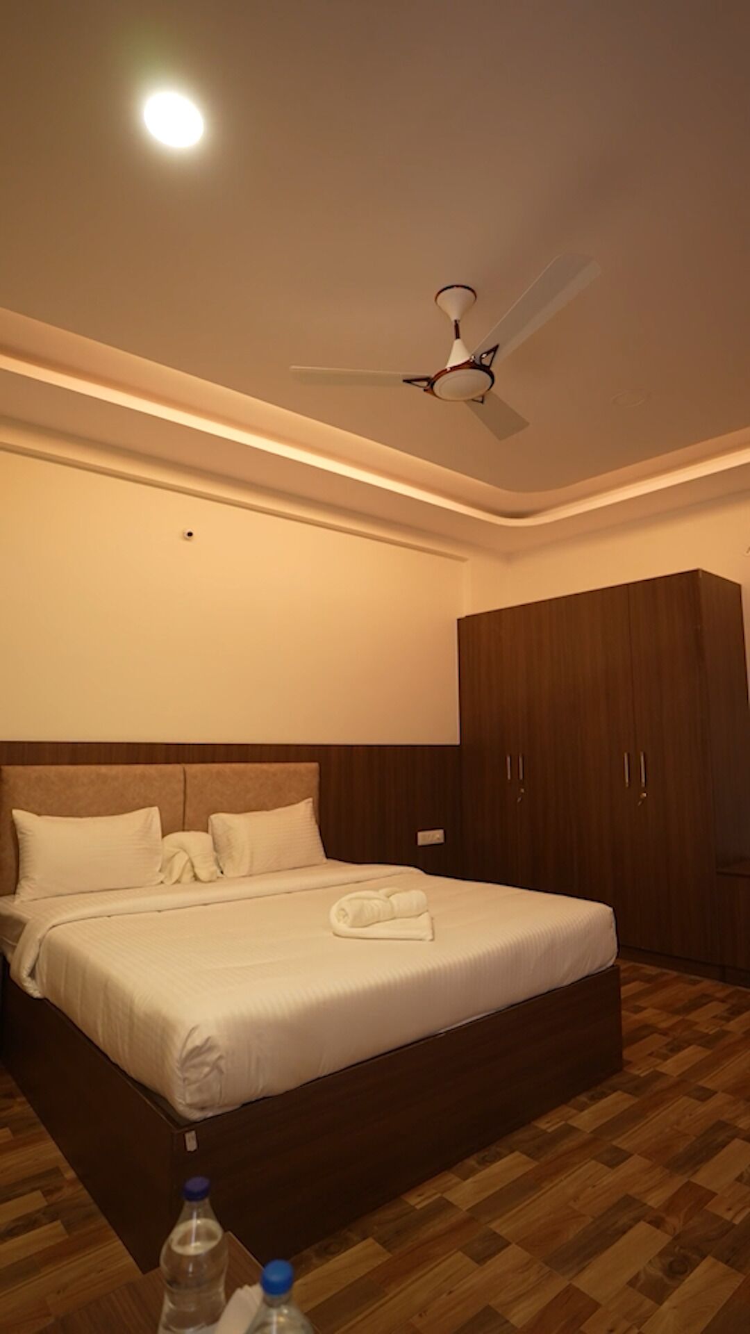 A hotel room with a large bed, a ceiling fan, wooden flooring, and a wardrobe.