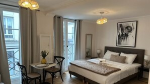Quarto de hotel com uma cama grande, uma mesa de jantar com cadeiras, uma pequena mesa com um vaso e uma varanda com cortinas.