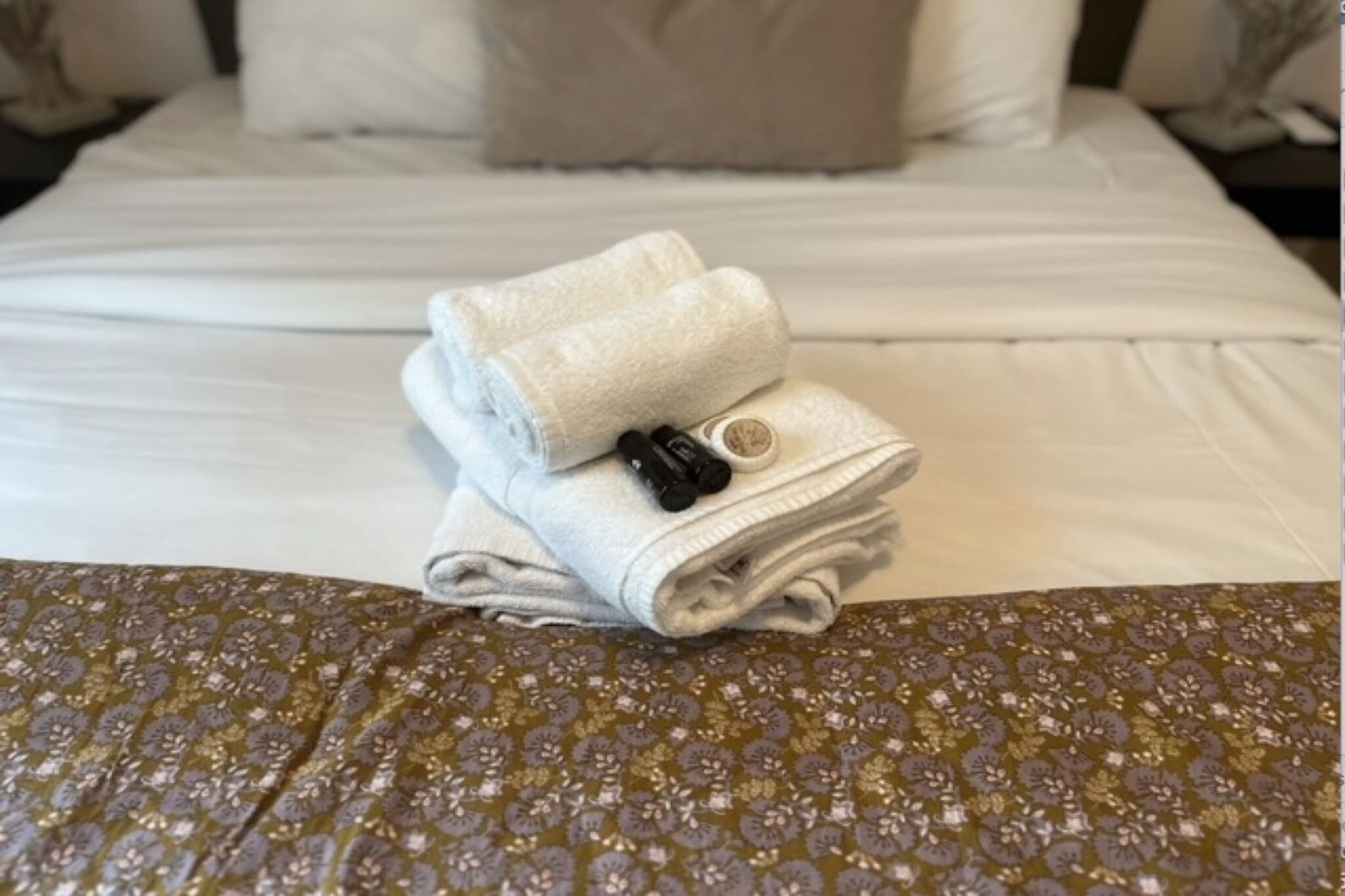 Una cama bien hecha con una toalla blanca doblada y dos gomas para el pelo negras encima.