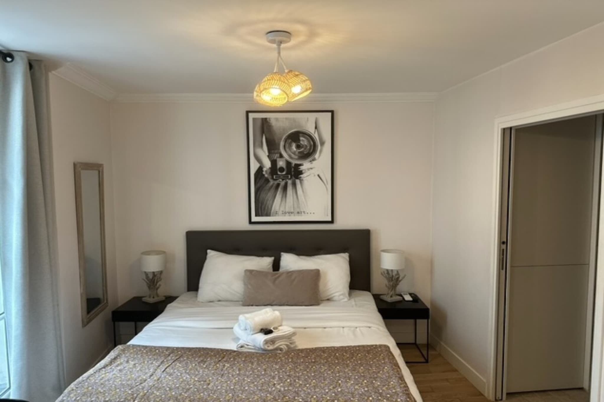 A bedroom with a bed, two bedside tables, a wall-mounted light fixture, a framed artwork, and a door.