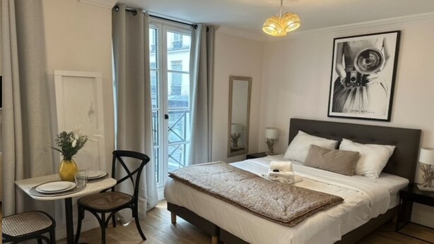 Quarto de hotel com cama, uma mesa de jantar com cadeiras, uma pequena mesa com um vaso e uma varanda com cortinas.
