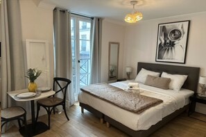 Een hotelkamer met een bed, een eettafel met stoelen, een kleine tafel met een vaas en een balkon met gordijnen.