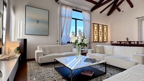A spacious living room with a large window, a white sofa, a coffee table, and a dining area with a table set for two. - Le Masque Terrace (Venecia)
