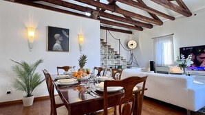 A dining area with a wooden table set for four, surrounded by chairs. There is a white sofa in the background, a television on the right, and a bookshelf with various items. - Le Masque Terrace (Venecia)