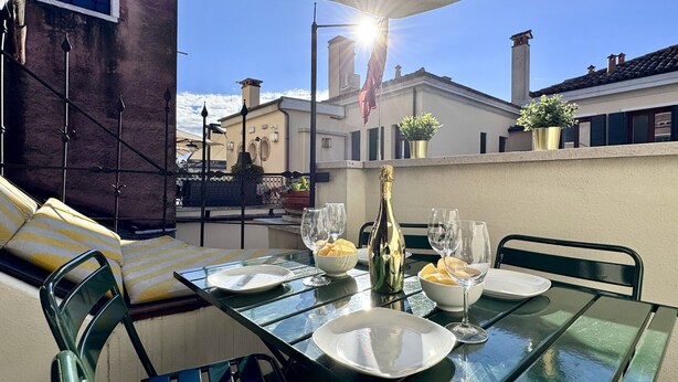 A rooftop terrace with a glass table set for two, featuring plates, glasses, and a bottle of champagne. - Le Masque Terrace (Venecia)