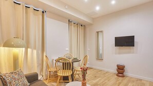 Un salon moderne comprenant un canapé, une table à manger avec des chaises, un téléviseur à écran plat fixé au mur et une petite table d’appoint avec un vase.