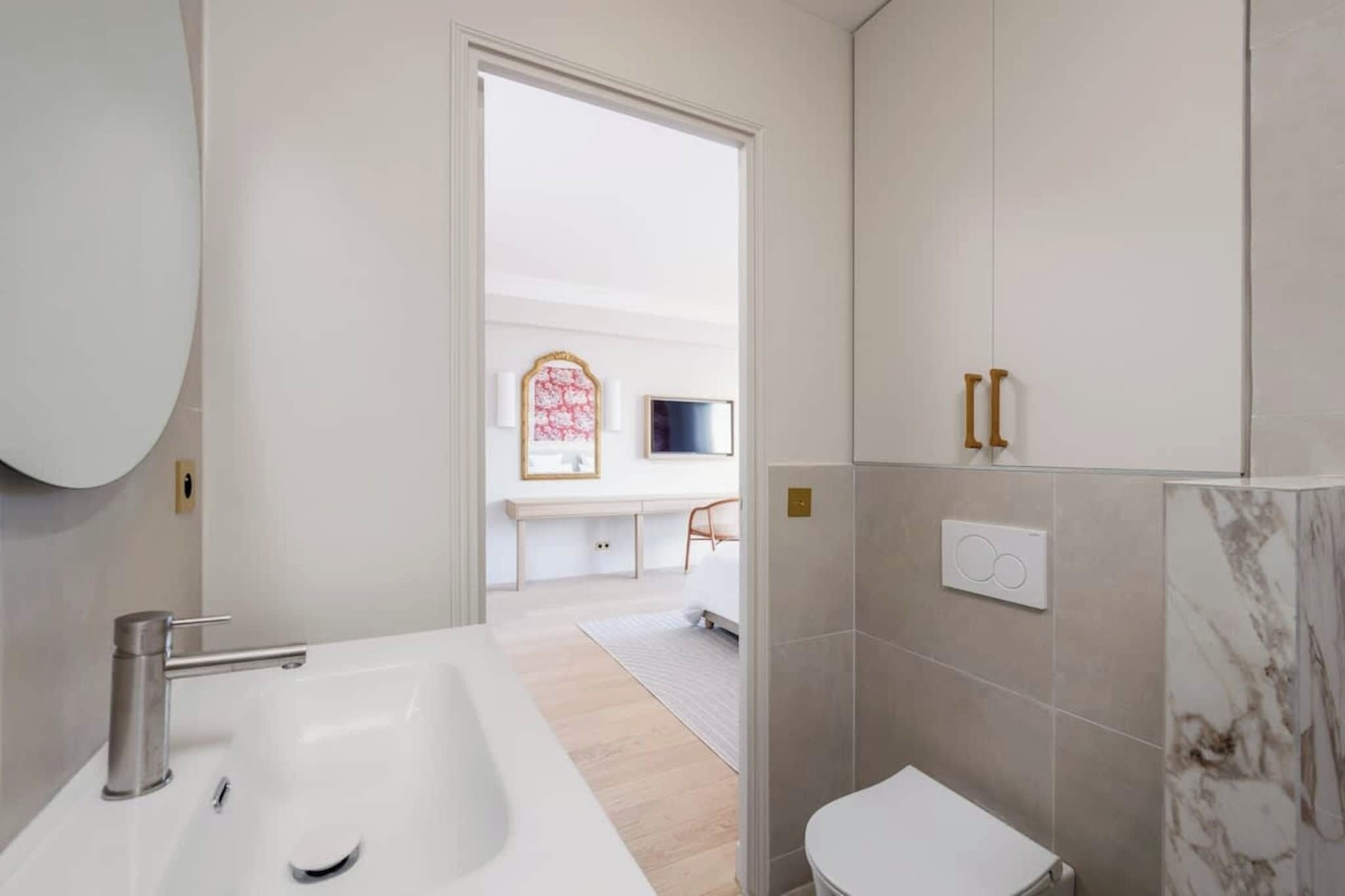 A modern bathroom with a large white sink, a toilet, and a shower area. There is a door leading to a bedroom with a bed and a TV.