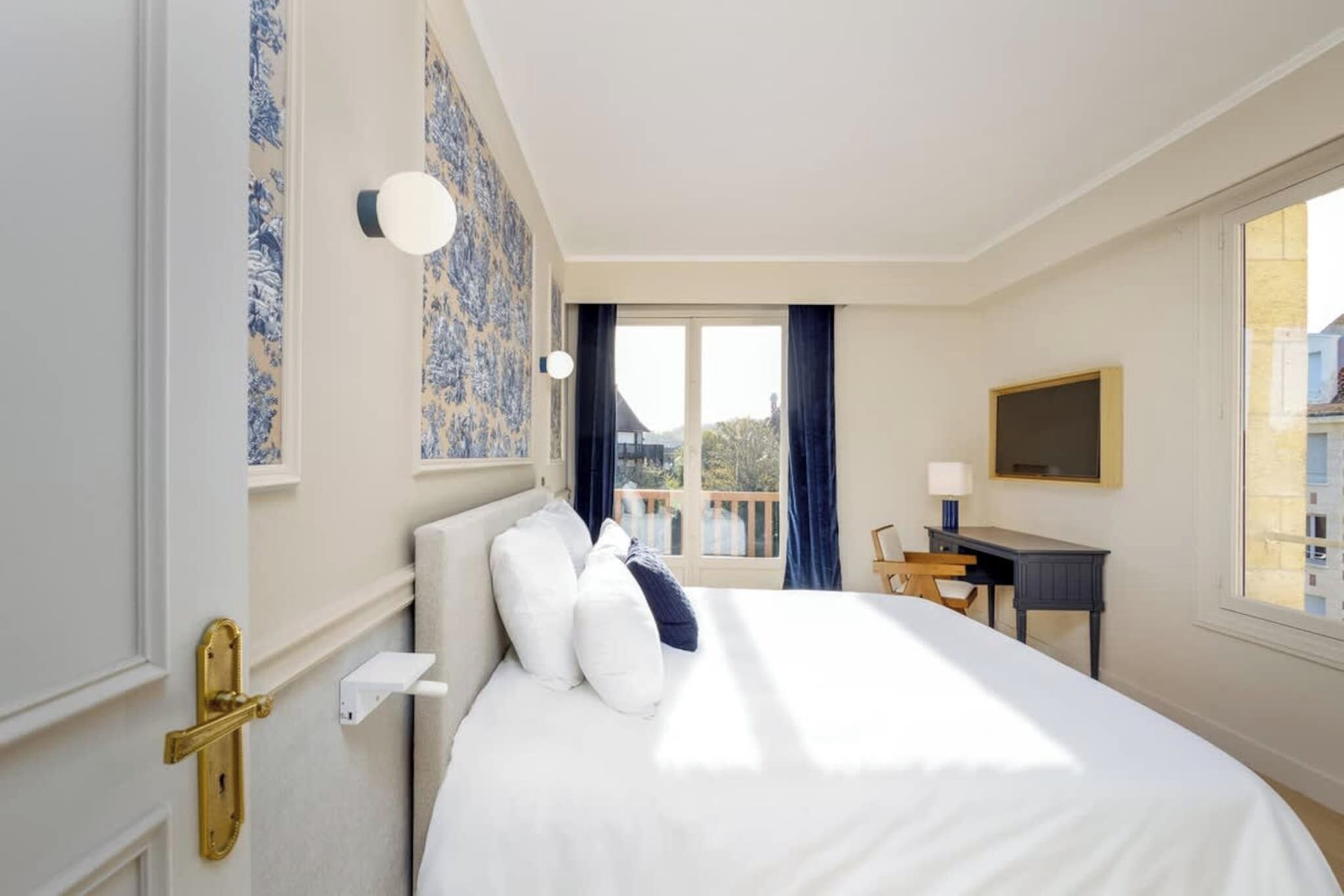 A hotel room with a bed, a desk, a chair, a TV, and a balcony view.