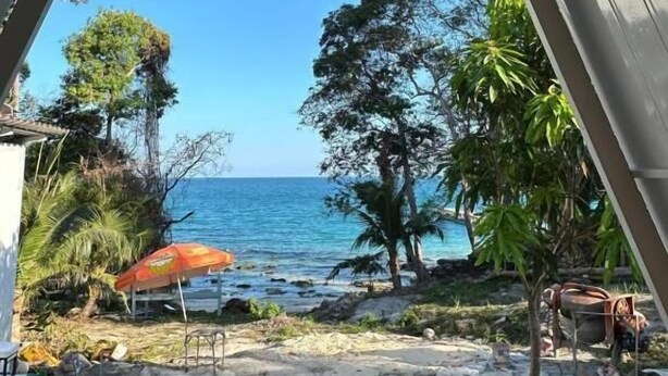 Blick von einer überdachten Außenfläche mit Sandboden, einem kleinen Pool und einem Strand in der Ferne.