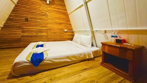 Ein kleiner, gemütlicher Raum mit holzverkleideter Schräge, einem Bett, einem Nachttisch und einem Wandventilator.