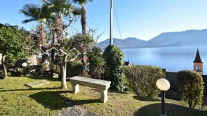 Um jardim com um banco de pedra, palmeiras e vista para um lago com montanhas ao fundo.
