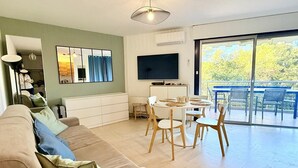 A modern living room with a sofa, a dining table with chairs, a flat-screen TV, and a large window with a view of trees. - Apartment for 4, pool, AC, WiFi, parking, 300m from the beach (Mandelieu-la-Napoule)