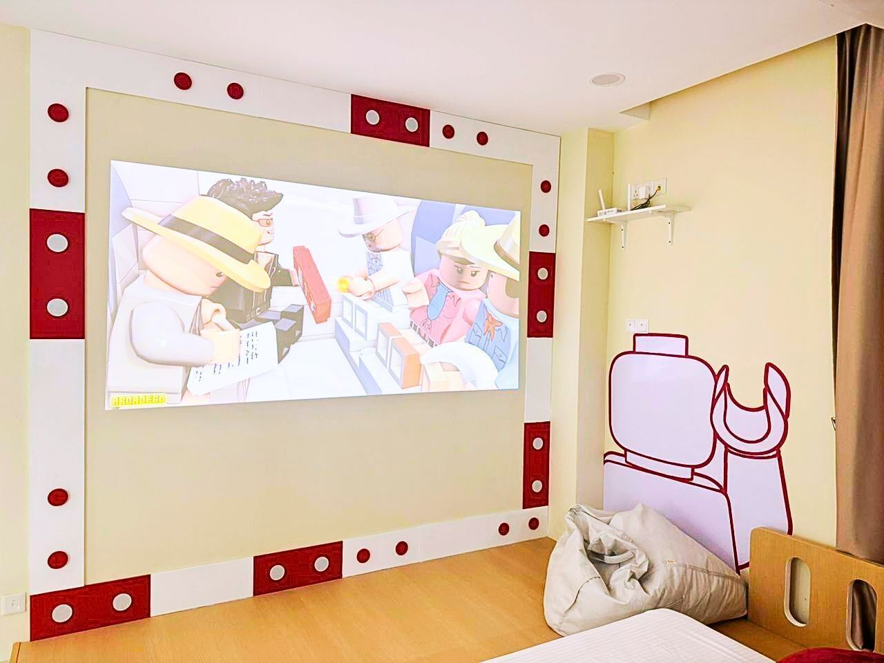 A hotel room with a television mounted on the wall, a bed with a pillow, a wooden headboard, and a wall-mounted shelf with a lamp.