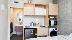 A compact hotel room with a kitchenette, a bed, a washing machine, and a small bathroom. - Petone Studio | Balcony + Full Kitchen (Lower Hutt)