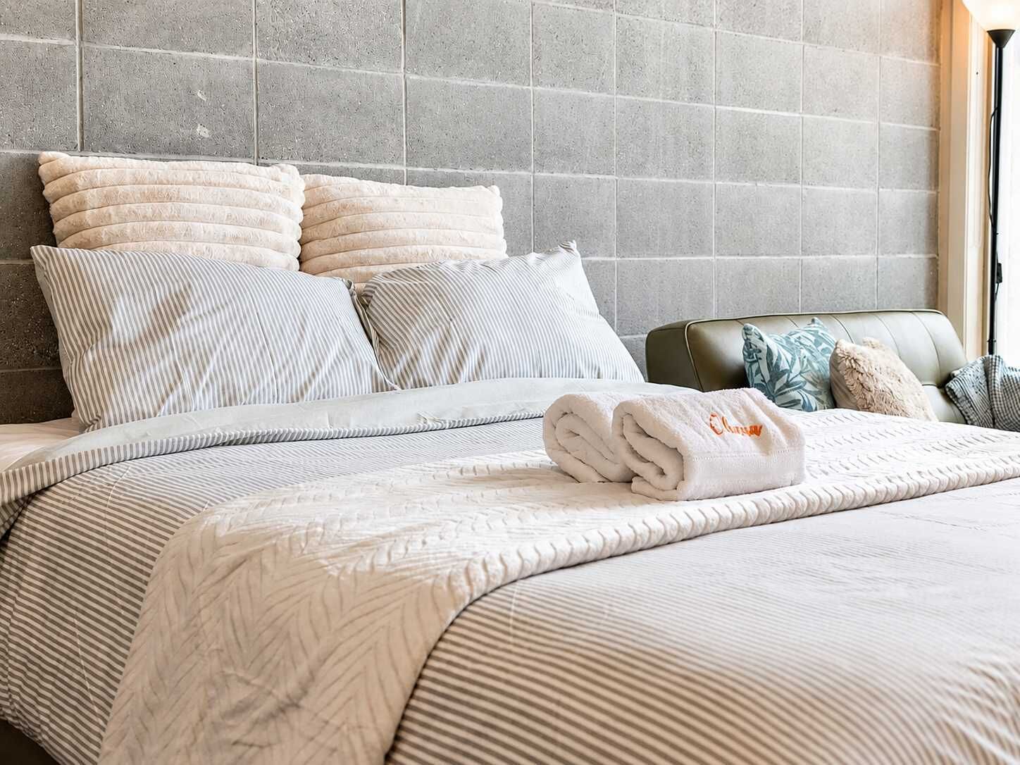 Un lit bien fait, avec une couvre-lit à rayures et des oreillers, une serviette pliée, et une tête de lit adossée à un mur carrelé.