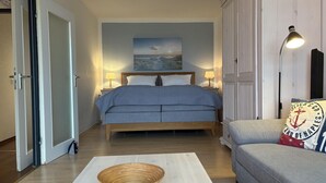 Una camera da letto con un letto, lampade sui comodini, un quadro, pavimento in legno, un divano grigio e un cesto su un tavolo.
