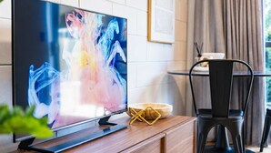 A modern living room with a flat-screen TV on a wooden console, a black chair, and a dining area in the background. - 1BR with Courtyard + Parking | Near CBD (Christchurch)