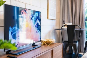 A modern living room with a flat-screen TV on a wooden console, a black chair, and a dining area in the background. - 1BR with Courtyard + Parking | Near CBD (Christchurch)