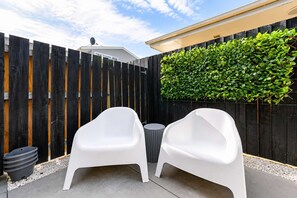 Un patio con dos sillas blancas, una mesa redonda pequeña y un cubo de basura negro.