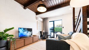 A modern living room with a flat-screen TV, wooden furniture, a dining area, and a staircase. - 1BR with Courtyard + Parking | Near CBD (Christchurch)