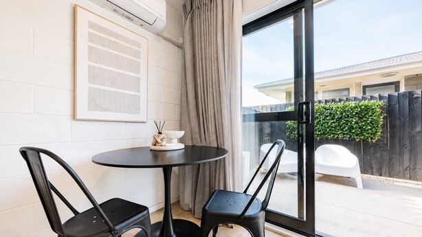 A room with a round table and two chairs, a framed artwork on the wall, and a sliding glass door leading to an outdoor area. - 1BR with Courtyard + Parking | Near CBD (Christchurch)