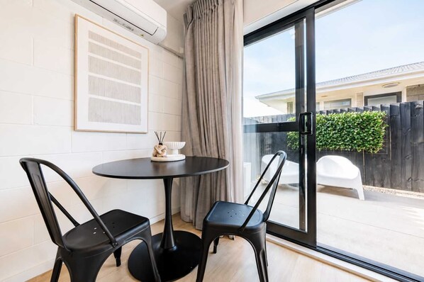 A room with a round table and two chairs, a framed artwork on the wall, and a sliding glass door leading to an outdoor area. - 1BR with Courtyard + Parking | Near CBD (Christchurch)