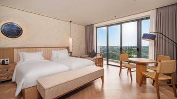A hotel room with a large bed, a desk, two chairs, a round table, a large window, and a view of the city.