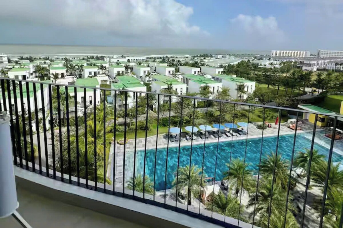 A balcony with a view of a swimming pool, a green lawn, and a distant body of water.