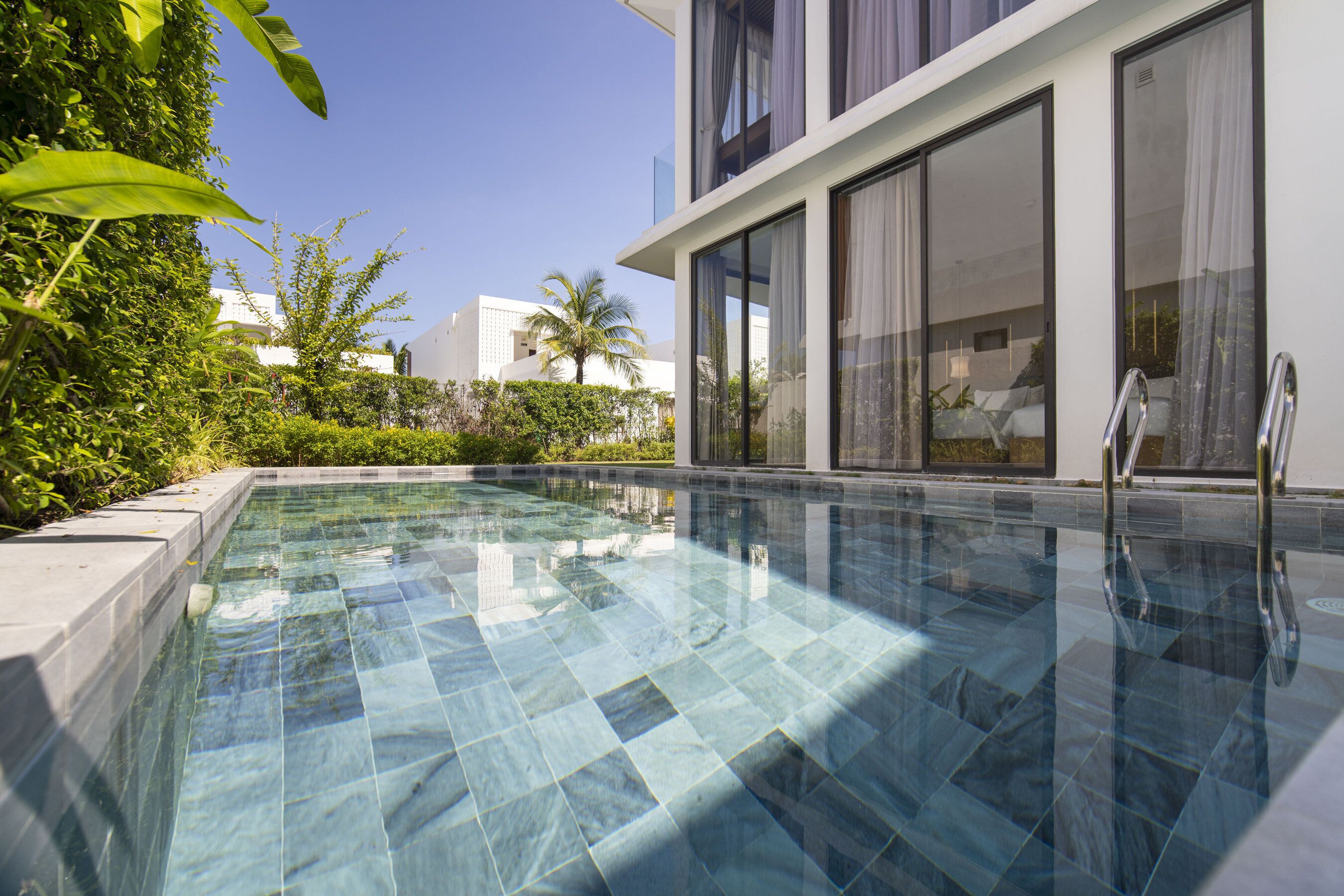 A modern pool area with clear water, surrounded by lush greenery and a contemporary building with large windows.