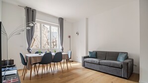 Un salon moderne avec un canapé gris, une table à manger avec des chaises et une grande fenêtre avec des rideaux.