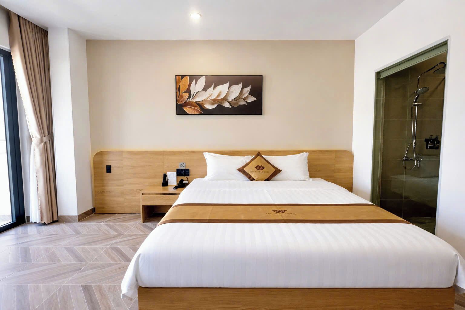 A hotel room with a large bed, a wooden headboard, a nightstand with a phone, a wall-mounted artwork, and a shower area.