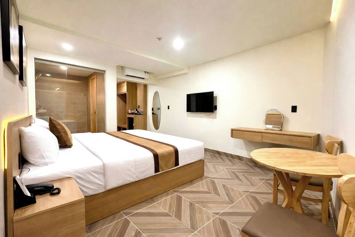 A hotel room with a bed, bedside table, desk, chair, TV, and a herringbone-patterned floor.