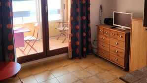 A room with a sliding glass door leading to an outdoor area, a wooden dresser with drawers, a small table, and a folding chair. - Large studio with view of Salses lake (Le Barcarès)