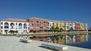 A row of colorful buildings with balconies, a reflective water body, and a paved walkway. - Large studio with view of Salses lake (Le Barcarès)