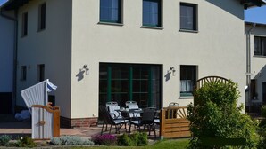 Kaksikerroksinen talo, jossa on patio, jossa on pöytä ja tuoleja, puiset portit ja pieni kukkapuutarha.